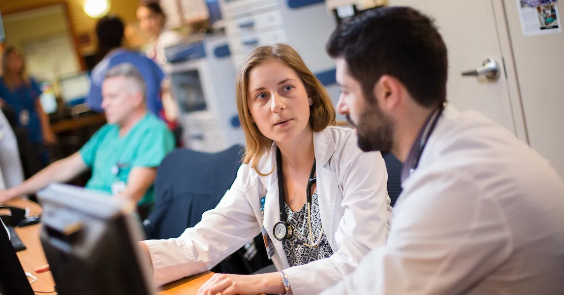 Two young medical students in white coats discussing a patient case at a hospital workstation. News