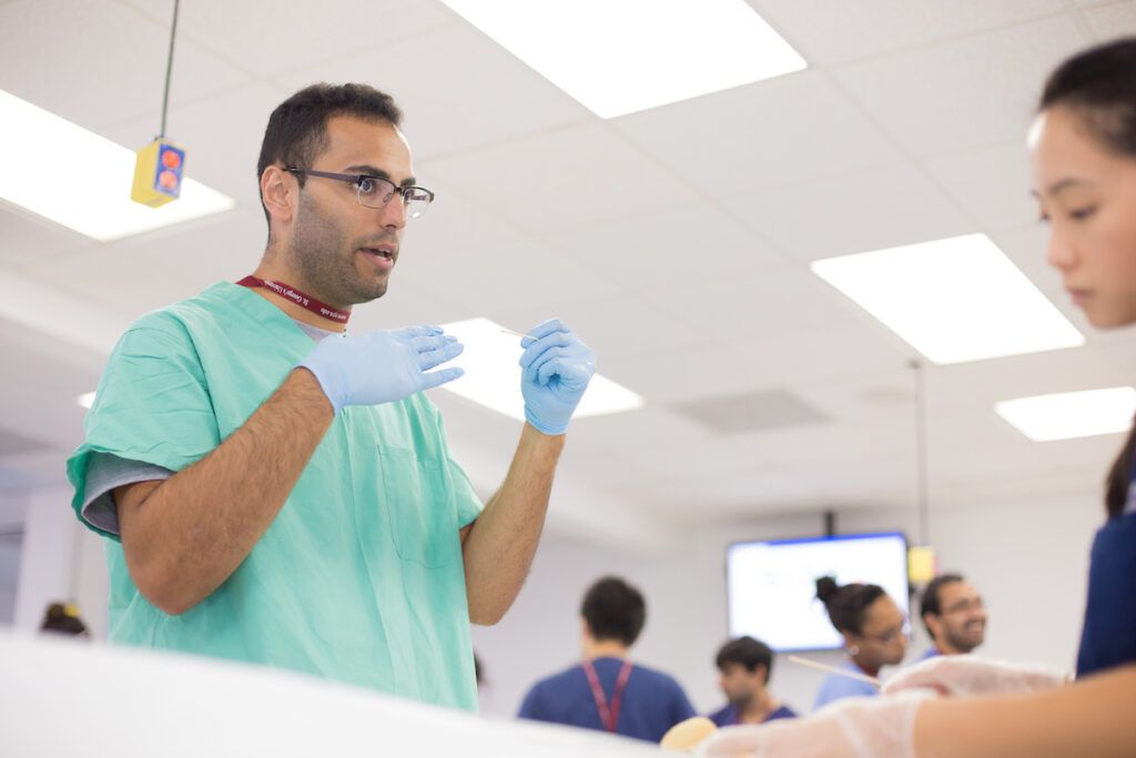 A medical student wearing green scrubs and blue gloves speaks and gestures during a laboratory class