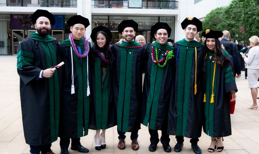 Seven graduates smiling together outdoors in black and green academic regalia News