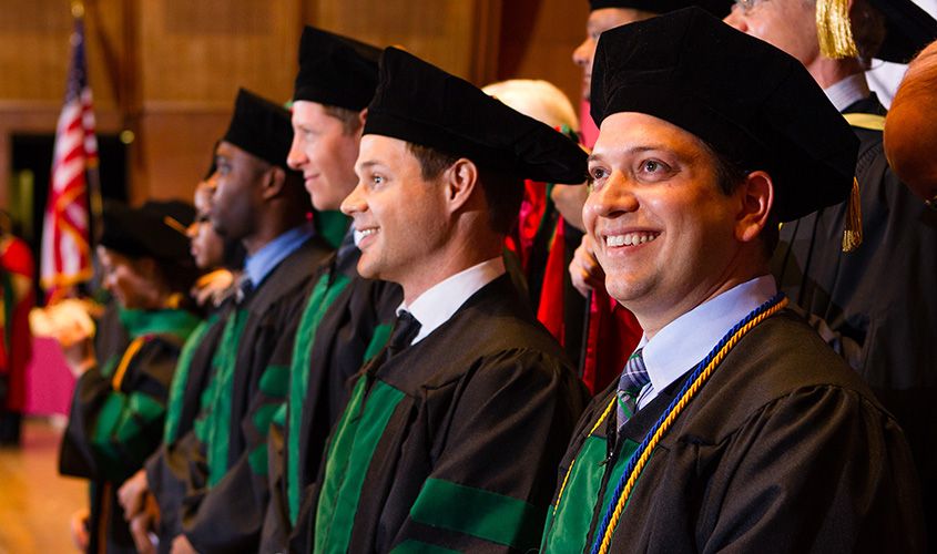 Proud SGU graduates in black and green academic regalia smile brightly at their medical school commencement ceremony.