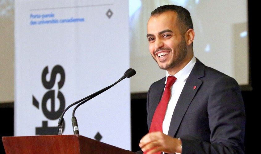 A smiling man in a suit and red tie speaks at a podium with microphones
