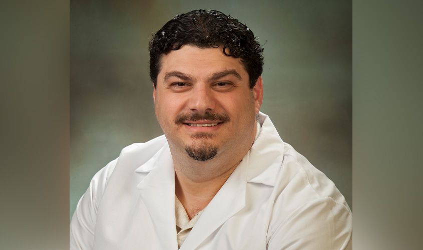 Smiling man with dark curly hair and a goatee wearing a white lab coat