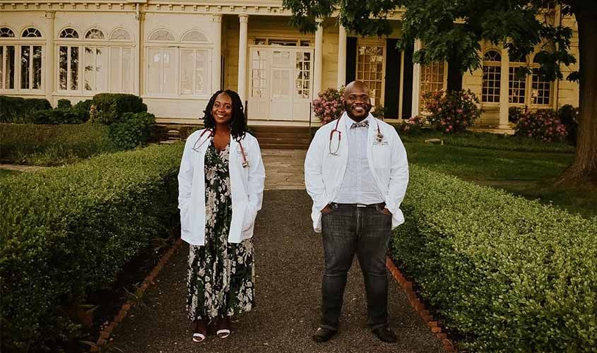Two doctors in white coats and stethoscopes smile on a hedge-lined pathway outside a historic building News
