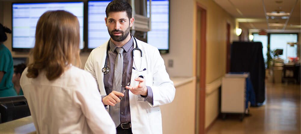 Male hospitalist speaks with another health care provider in hallway.