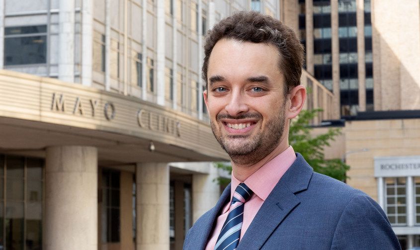 A smiling man in a blue suit and pink shirt standing outside the Mayo Clinic building entrance