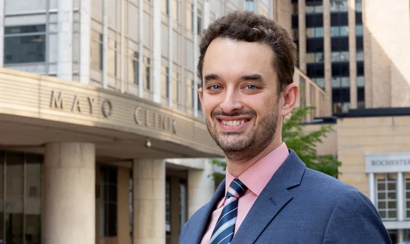 A smiling man in a blue suit and pink shirt standing outside the Mayo Clinic building entrance News