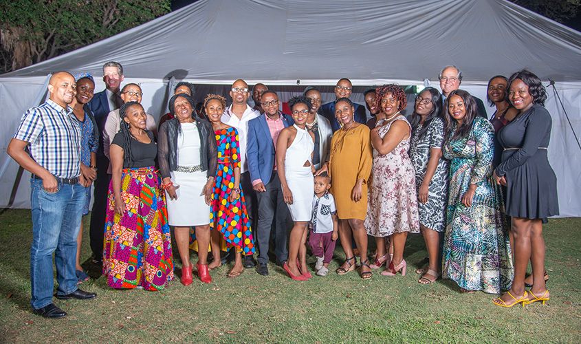 A large group of smiling people dressed in semi-formal attire posing outdoors in front of a white tent