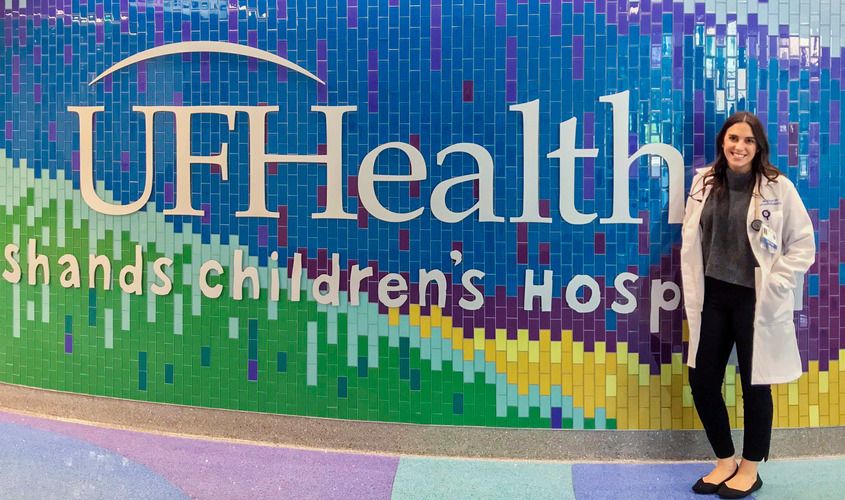 A doctor in a lab coat stands smiling in front of a colorful mosaic wall at UF Health Shands Children's Hospital News