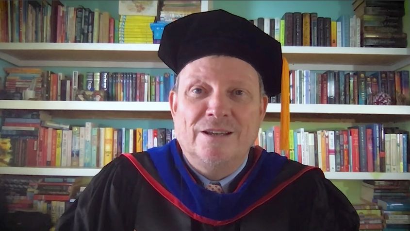 A smiling man wearing academic regalia with a blue and red hood in front of bookshelves News