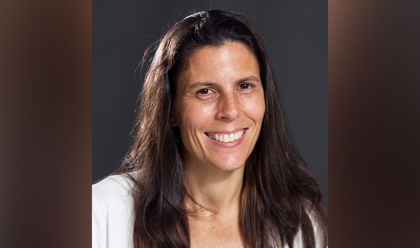 Smiling woman with long brown hair wearing a white top against a dark background