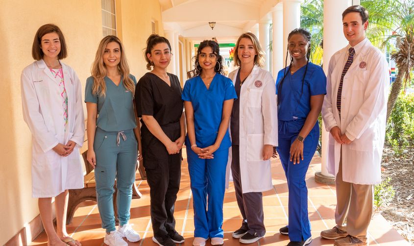 Seven healthcare students and professionals smiling in a row wearing scrubs and white lab coats in an outdoor hallway News