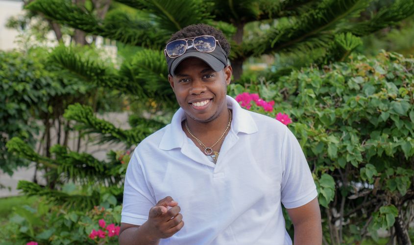 Smiling man in a white polo shirt and visor pointing toward the camera in front of lush garden greenery News