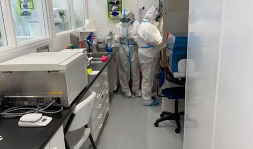 Two laboratory researchers wearing full white personal protective equipment (PPE) working with scientific equipment in a compact testing facility