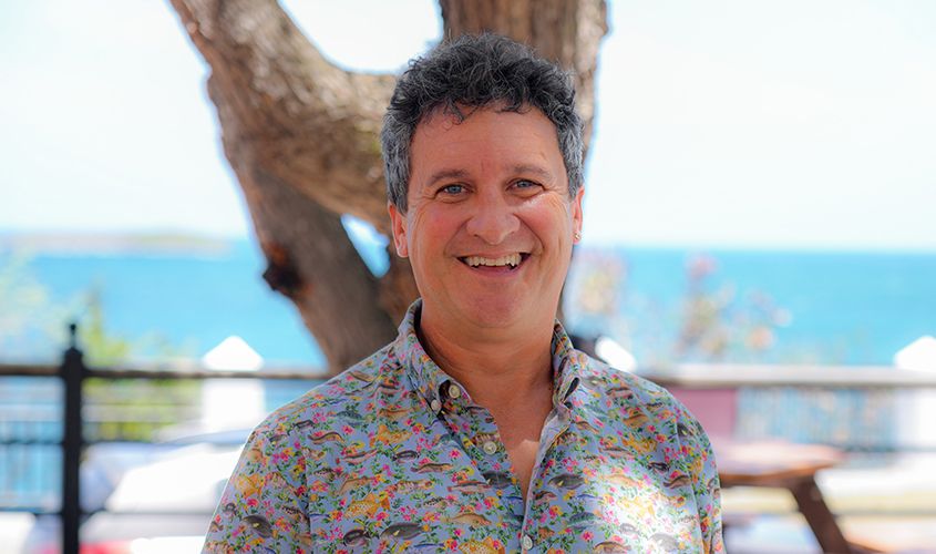 Smiling man wearing a colorful fish-patterned shirt, standing outdoors with an ocean view in the background News