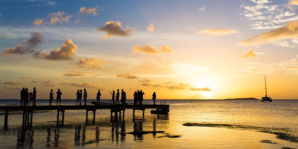 Silhouettes of students and families on a wooden pier watching a beautiful golden sunset over the water in Grenada during SGU Family Weekend.