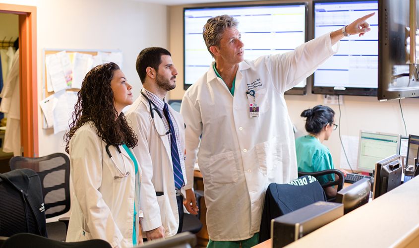 A physician points to a wall-mounted display while discussing information with two colleagues in a hospital News