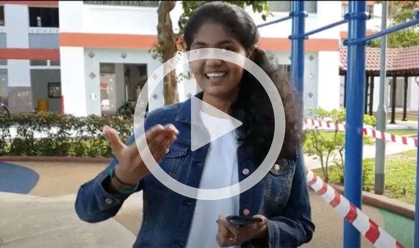 Smiling young woman in a denim jacket speaking and gesturing outdoors, with a play button overlay News