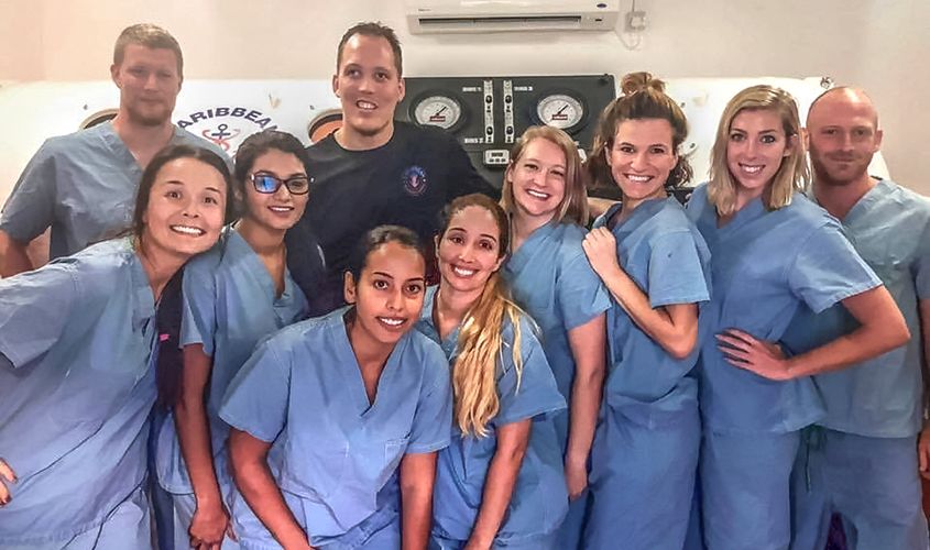A diverse group of SGU medical students and faculty in blue scrubs posing together in front of a hyperbaric chamber on the Grenada campus. News