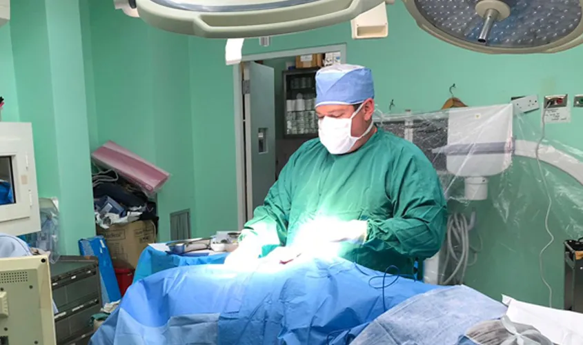 A surgeon in green scrubs and a mask operates on a patient covered in blue drapes in an operating room