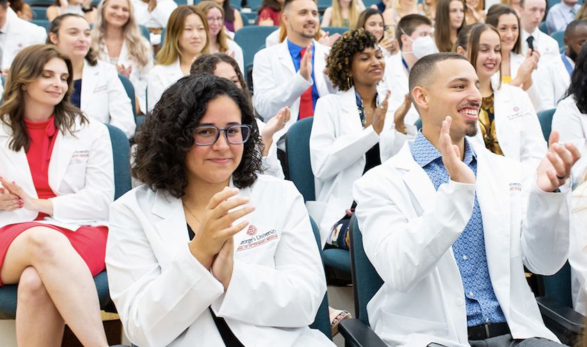 Students in white coats smile and applaud during a university white coat ceremony in an auditorium News