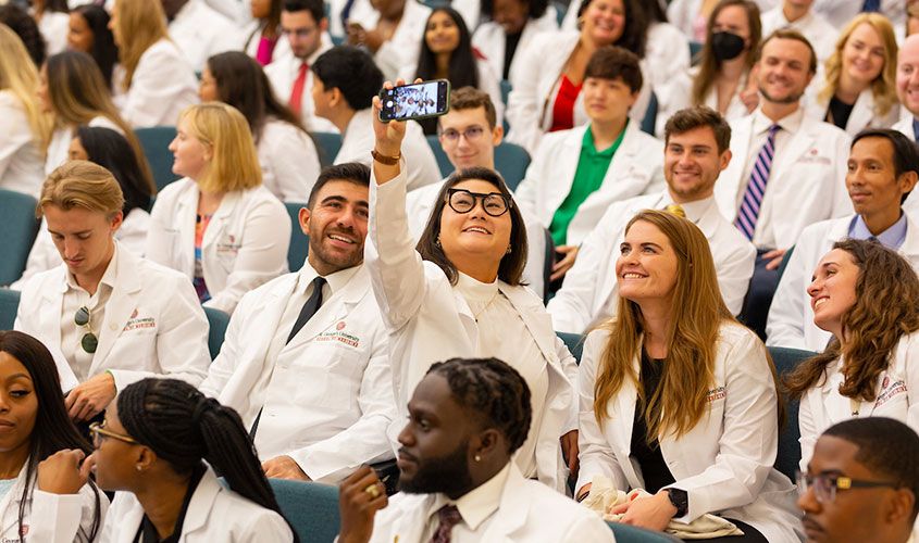 Medical students in white coats smile as one takes a selfie in a crowded lecture hall News