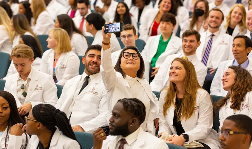 Medical students in white coats smile as one takes a selfie in a crowded lecture hall