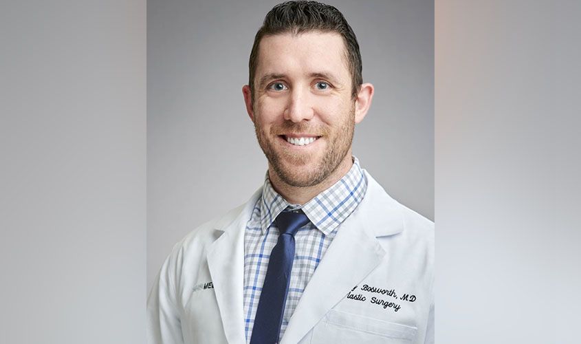 Smiling doctor wearing a white lab coat embroidered with Plastic Surgery, a blue tie, and a plaid shirt News