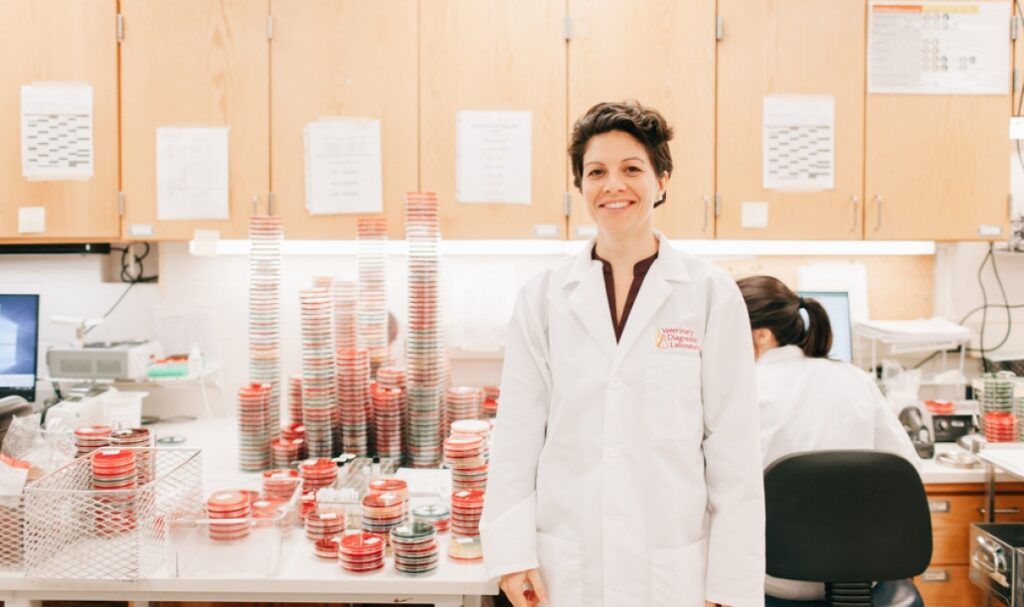 Smiling scientist in a white lab coat standing beside tall stacks of petri dishes in a diagnostic laboratory