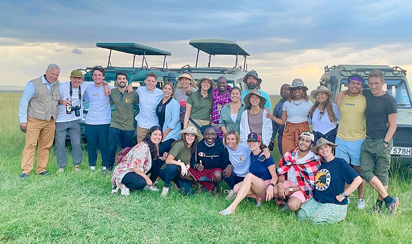 A large group of tourists and safari guides pose together in a grassy field in front of green safari vehicles News