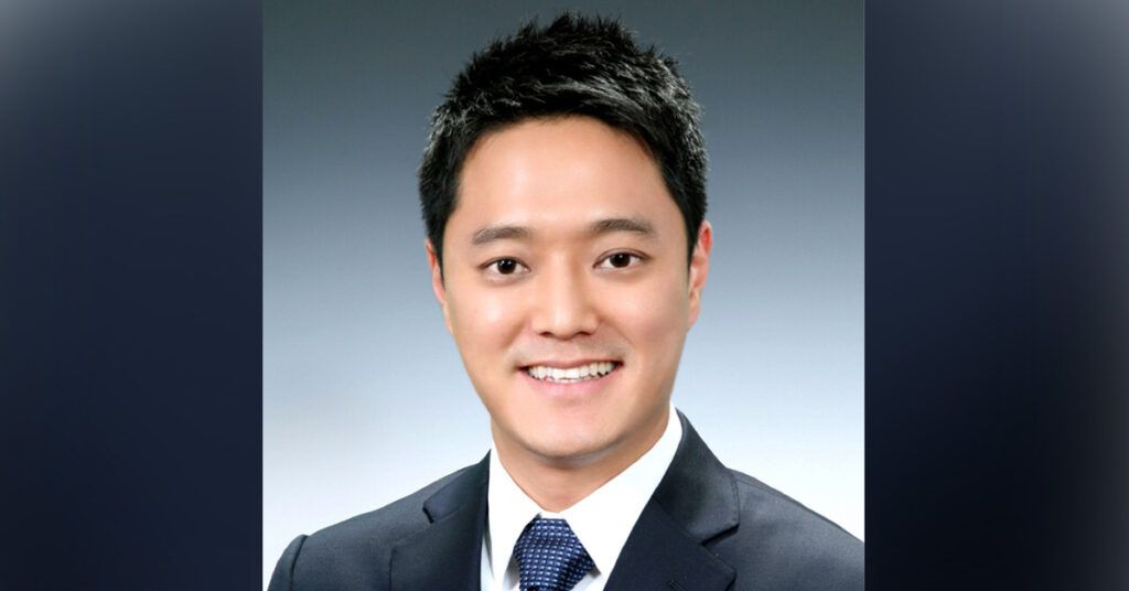 Smiling man with short dark hair wearing a dark suit, white dress shirt, and blue patterned tie