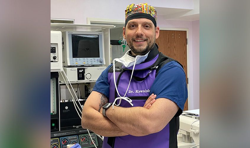 Smiling doctor named Dr. Kowalski wearing surgical scrubs and a protective apron in front of medical equipment