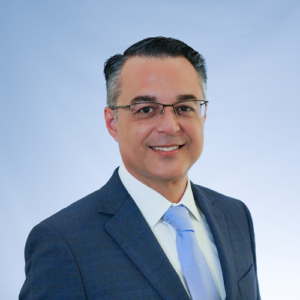 Smiling man with glasses wearing a dark suit jacket, white collared shirt, and light blue tie