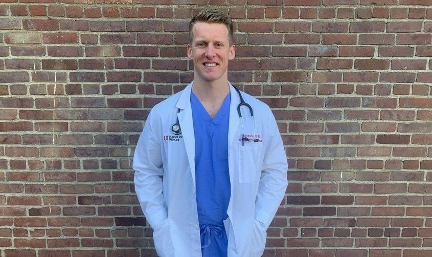 Smiling doctor wearing blue scrubs, a white lab coat, and a stethoscope, standing against a brick wall