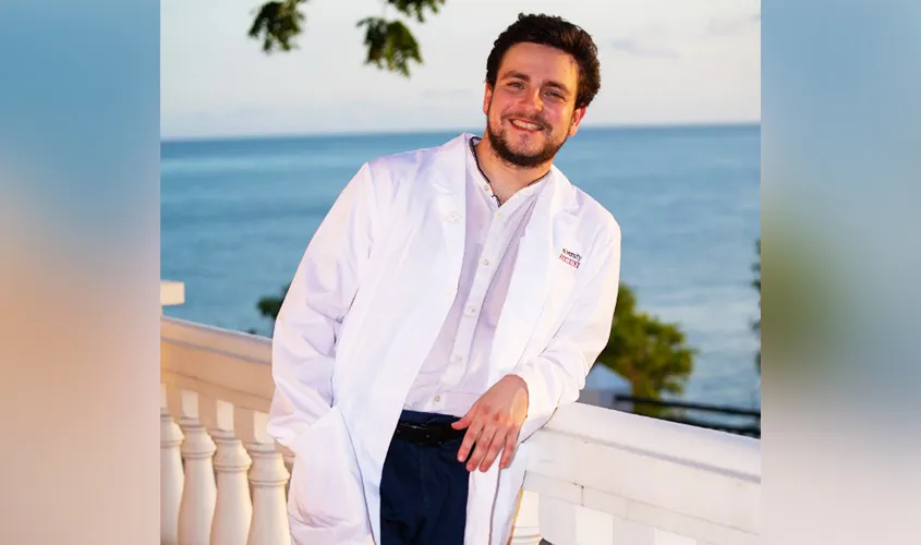 Smiling man in a white lab coat leaning against a balcony railing overlooking the ocean