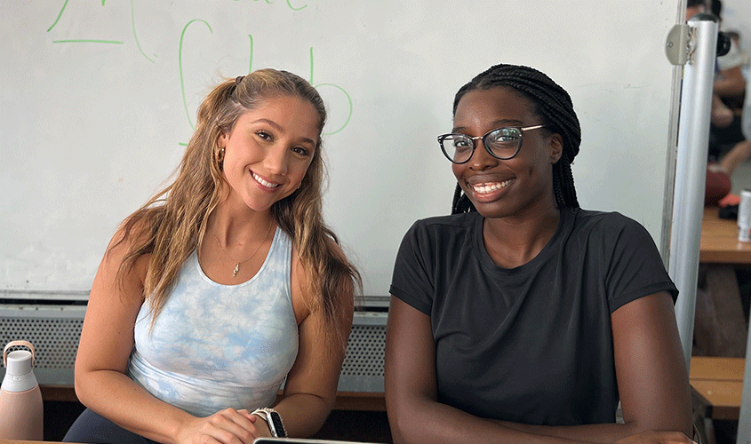 Two smiling women sit side by side in front of a whiteboard in a classroom