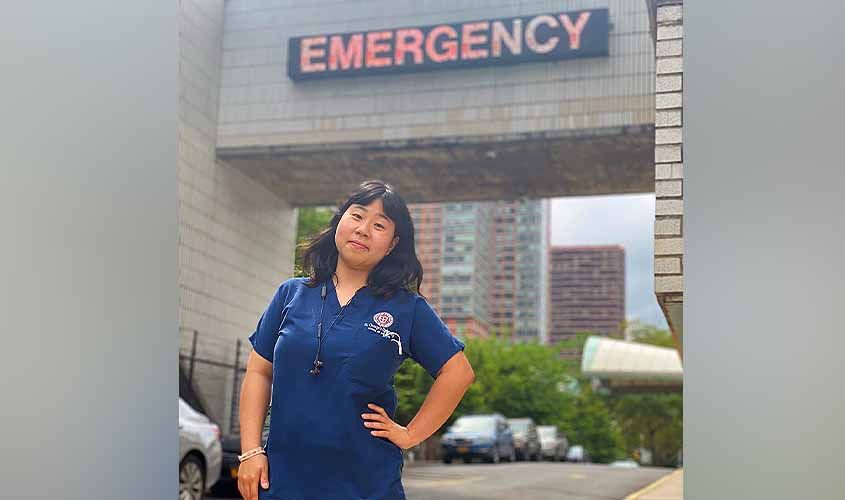 A healthcare worker in blue scrubs stands smiling beneath an illuminated Emergency entrance sign outside a hospital