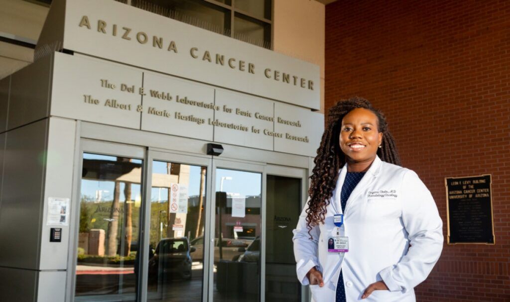 A doctor wearing a white lab coat smiles in front of the entrance to the Arizona Cancer Center