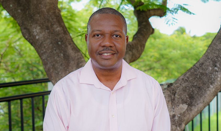 A smiling man in a light pink collared shirt standing outdoors in front of leafy green trees News