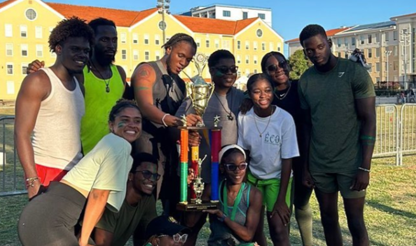 A group of smiling young adults poses outdoors while holding a large championship trophy