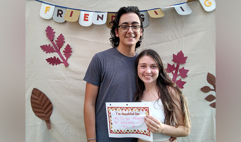 Couple at a Friendsgiving event holding a sign reading, "I'm thankful for My S.O. for moving to Grenada w/ me News