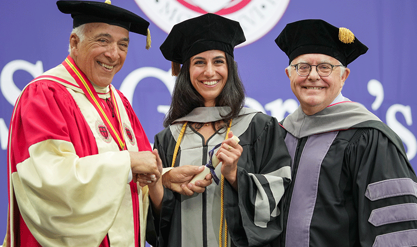 A graduate smiles while receiving her diploma and shaking hands with university officials during commencement