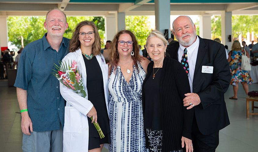 A medical student in a white coat holds flowers while posing with family members at a ceremony