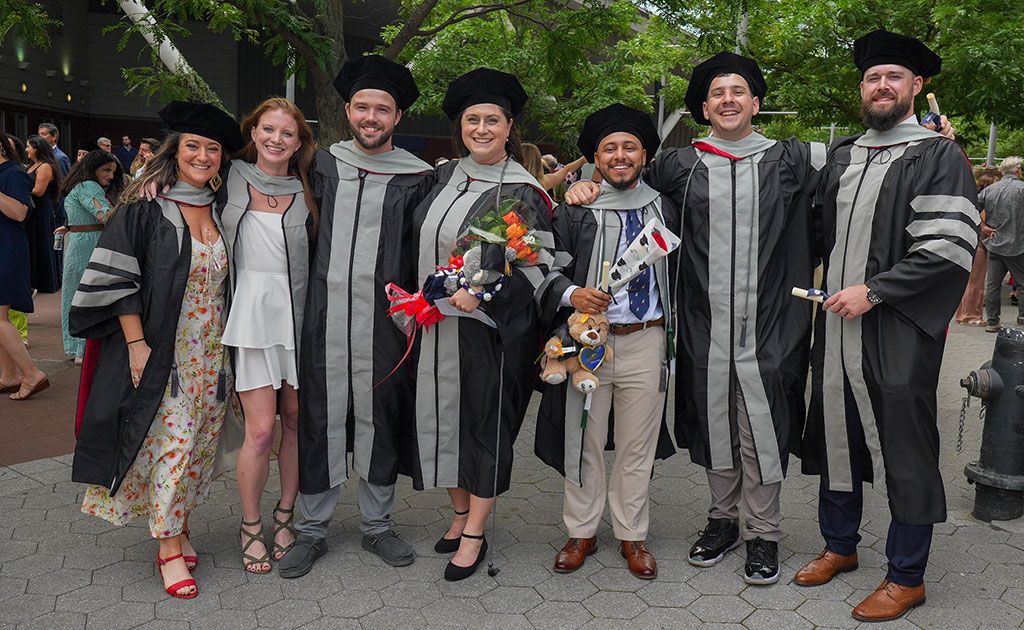 Seven smiling graduates posing together outdoors in academic regalia, holding diplomas and flowers