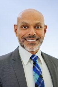 Smiling bald man with a salt-and-pepper beard wearing a grey suit and blue plaid tie