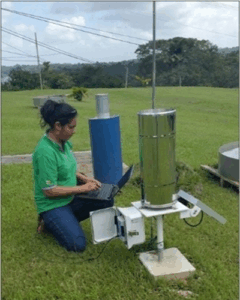 A woman uses a laptop connected to an outdoor weather station rain gauge in a grassy field