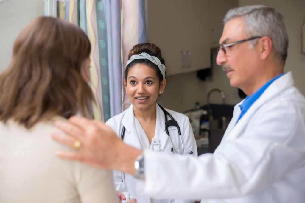 Female medical student on a clinical rotation observing a senior physician consulting with a female patient in an examination room.