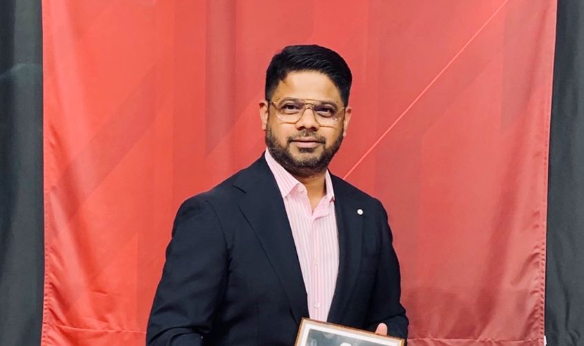 Man in a blazer and glasses holding a framed award in front of a red backdrop