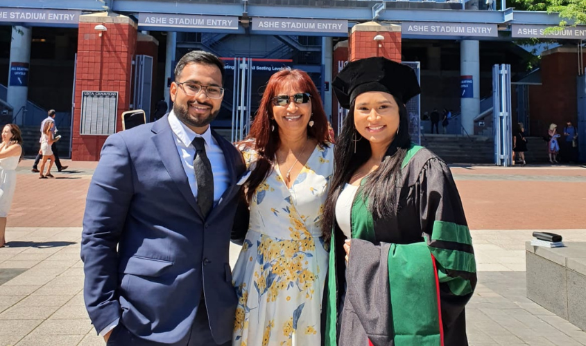 A graduate in an academic cap and gown smiling with two family members outside an Ashe Stadium entrance