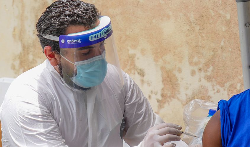A healthcare worker in full white PPE and a face shield administers a vaccine via injection at a community clinic.