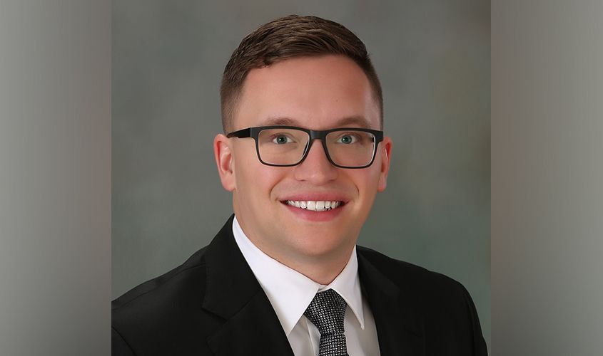 Smiling man with black glasses wearing a suit, white shirt, and tie against a studio background News
